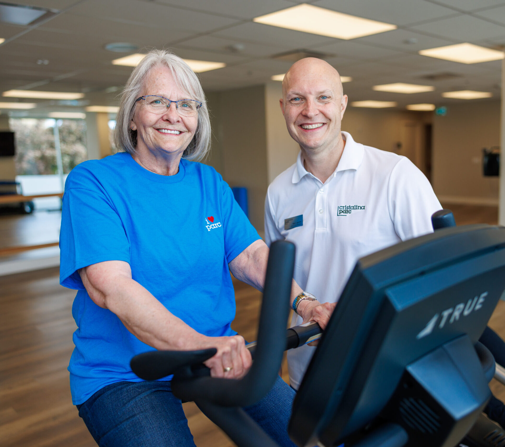 A senior resident on an exercise bike at the gym, the fitness trainer standing next to her. Independent Retirement Community.